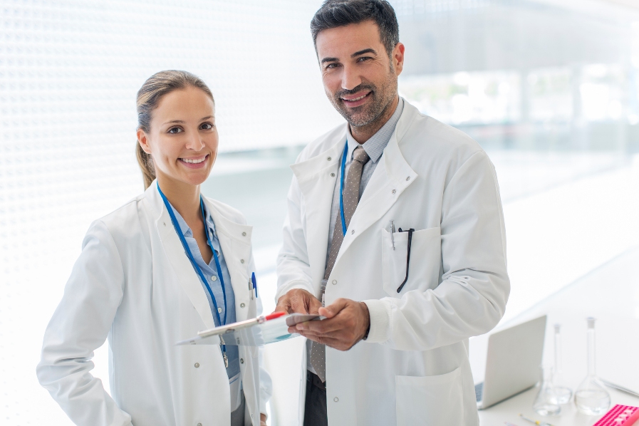 Una médica y un médico sonríen mirando hacia el frente mientras el médico sostiene un formulario.