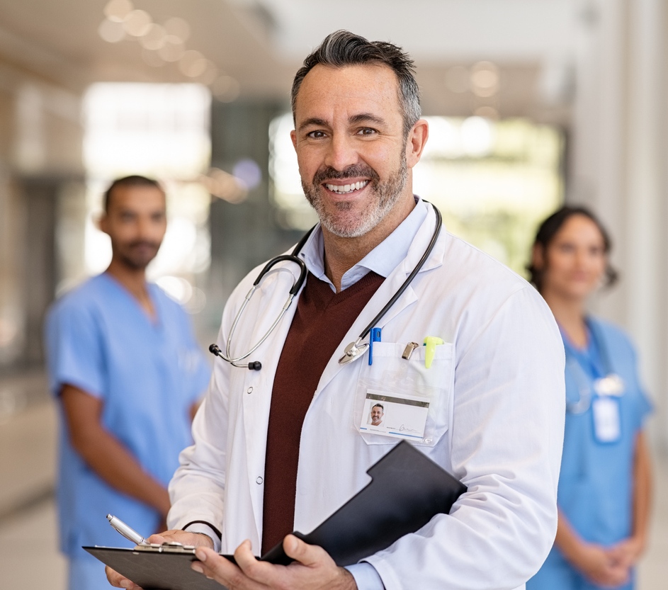 En el primer plano se ve un médico sonriente, en el segundo plano se ven un hombre y una mujer profesionales de la salud que también sonríen. Todos se encuentran en un pasillo de una clínica.