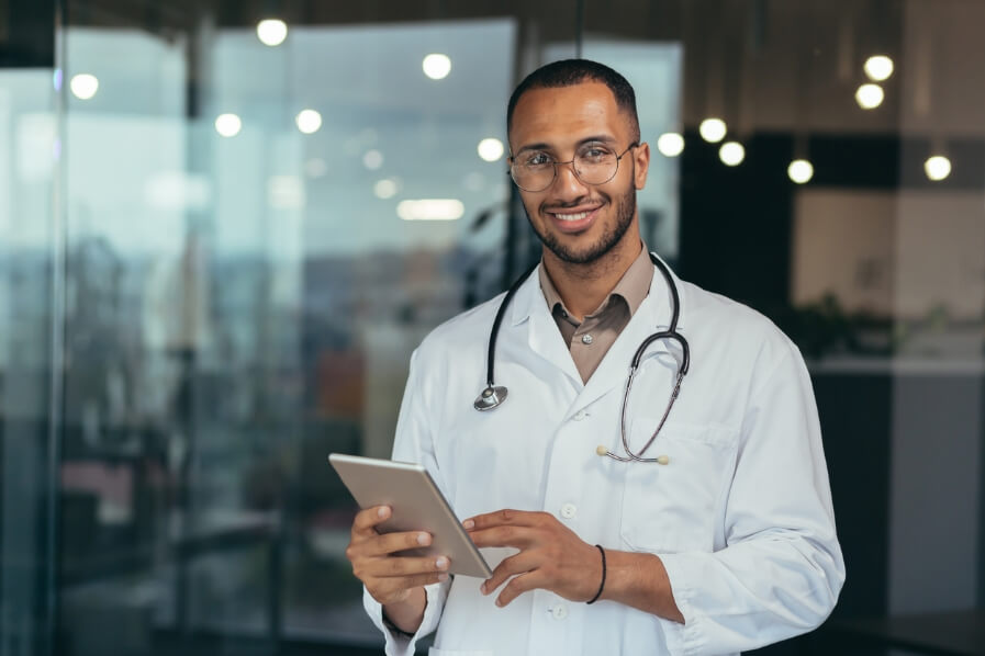 Hay un médico sonriendo mientras sostiene una tablet, al fondo se puede ver una recepción de una clínica.