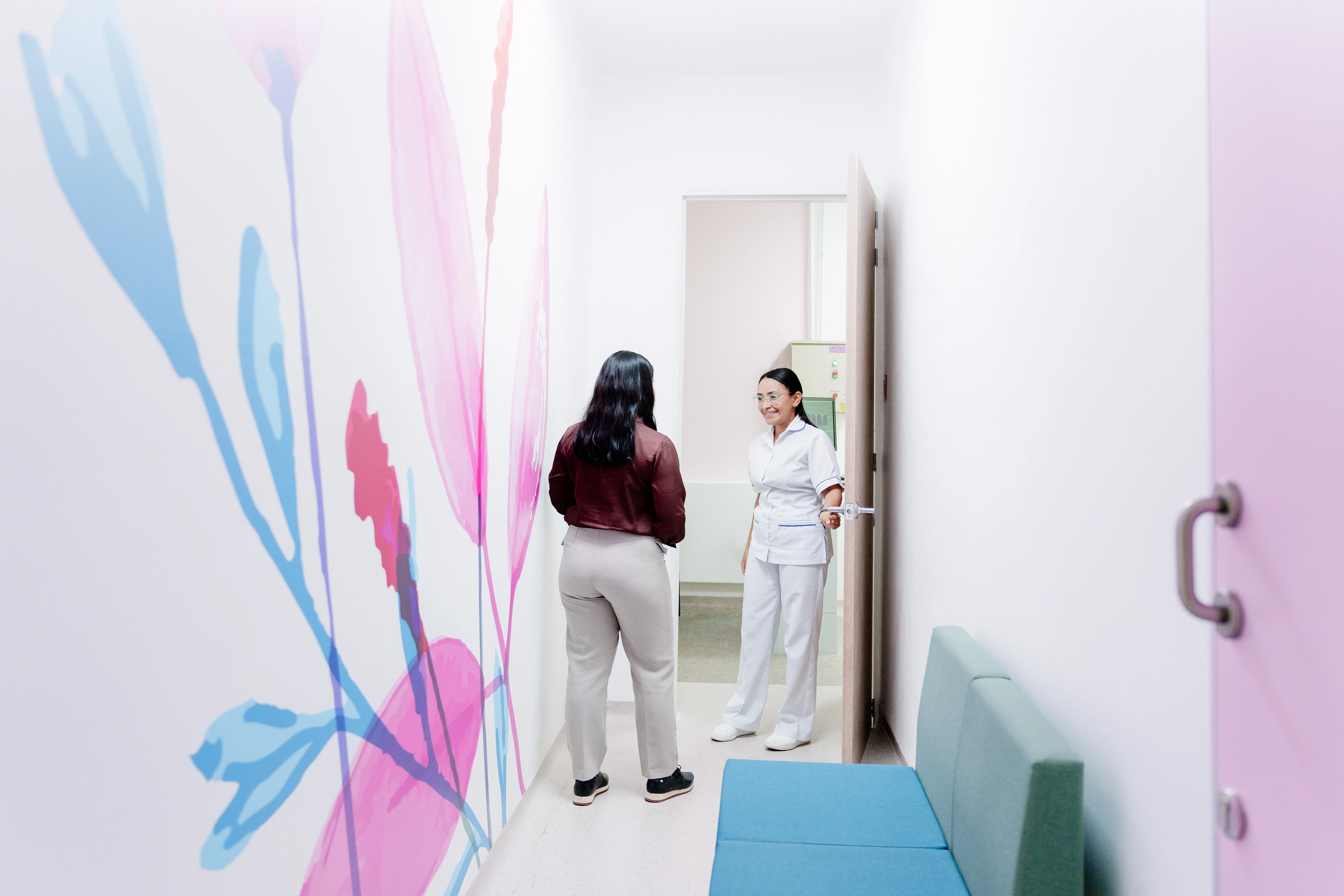 Hay un pasillo que en su pared izquierda tiene un mural de flores, al final hay una puerta en la cual una profesional de la salud le dala bienvenida a una paciente.