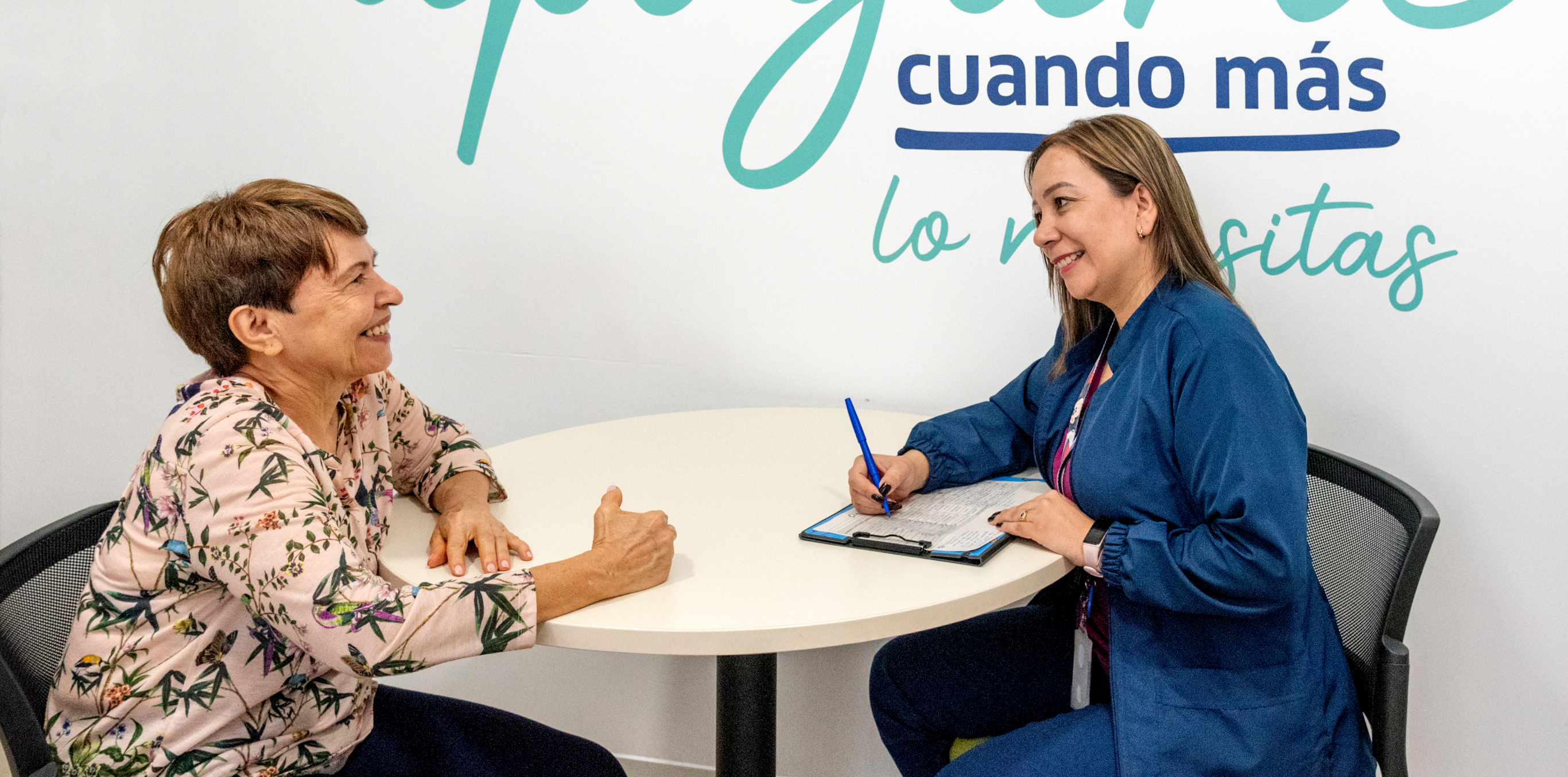 Profesional de la salud sentada en un escritorio, diligenciando a mano una planilla, con los datos que le proporciona la paciente que tiene enfrente.